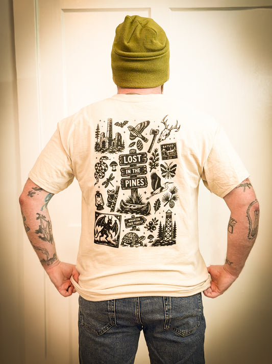 A man viewed from behind wearing a cream-colored t-shirt with a large black graphic print on the back, paired with a green beanie and blue jeans, standing in front of a white door.