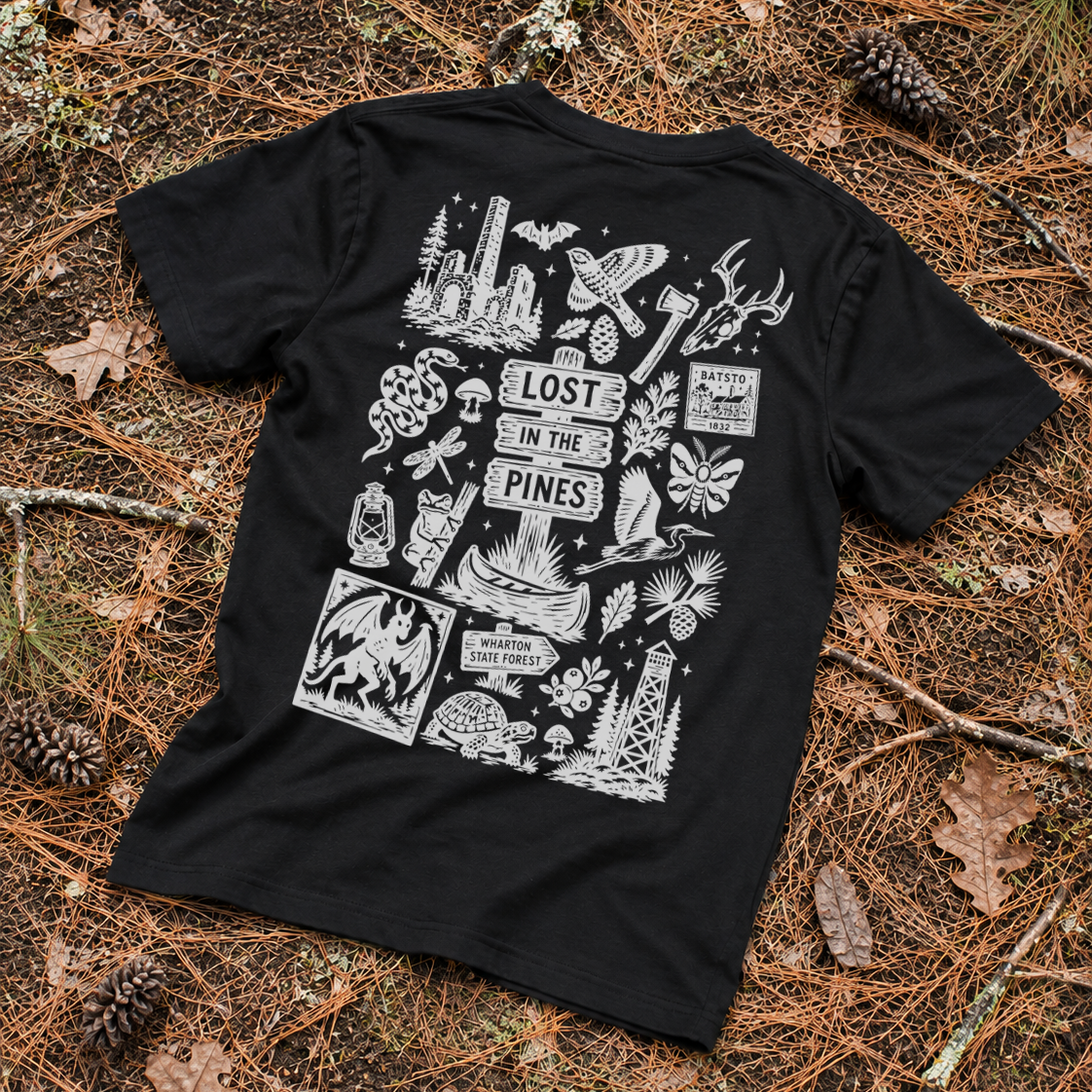 A black short-sleeve T-shirt laid flat on a forest floor covered in pine needles and pinecones, showing a large white illustrated back graphic with the words 'LOST IN THE PINES' and various outdoor-themed drawings.
