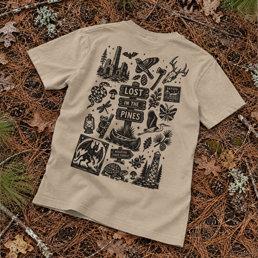 A beige graphic t-shirt laid flat on a forest floor covered in pine needles and leaves. The back of the t-shirt features a large black graphic print with various nature and survival-themed illustrations, including a snake, axe, antler, and a boat, surrounding the text 'LOST IN THE PINES'.